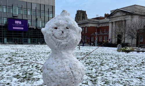 Large snowman with a smiley face made of small stones, standing on a snow-covered lawn in front of modern and historic buildings.